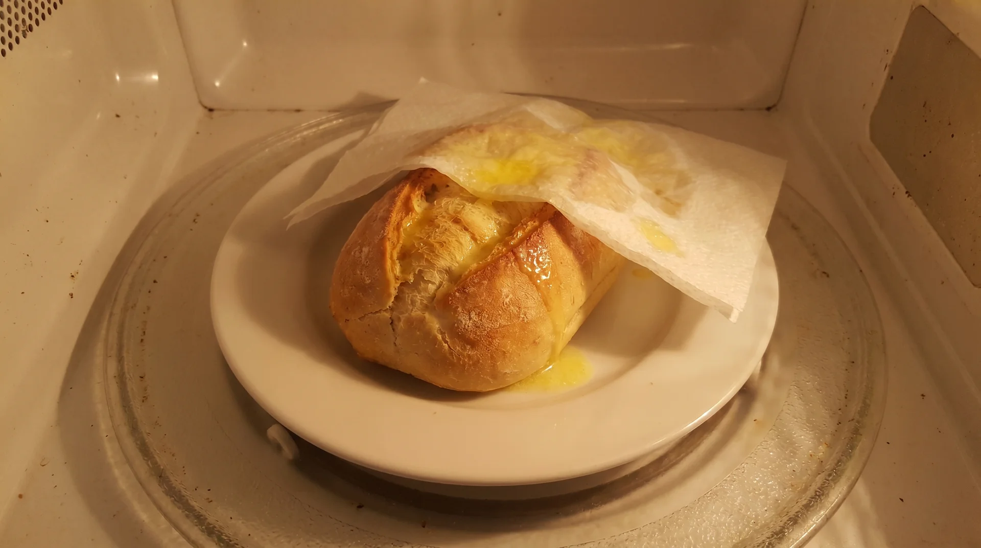 Butter-coated bread roll covered with a plain white paper napkin on a microwave plate, illustrating the fat-contact fire risk
