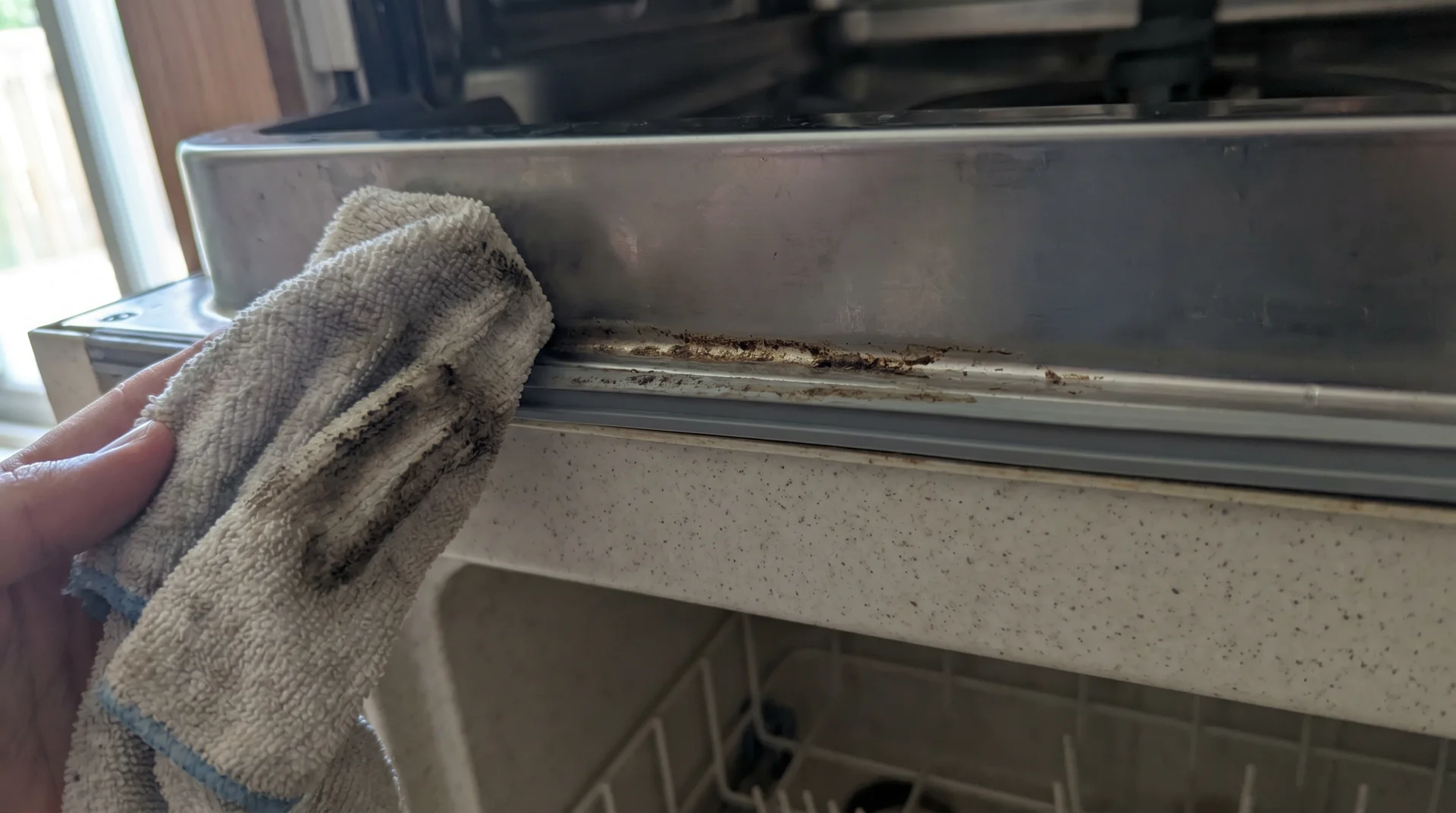 Cloth wiping the debris-collecting bottom edge of a dishwasher door interior, commonly missed during cleaning