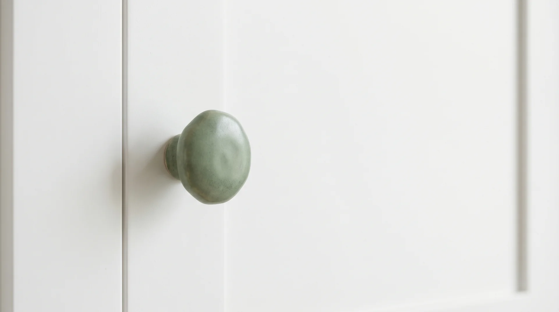 Handmade sage green ceramic knob on white shaker cabinet door — the single color in an otherwise all-white frame 