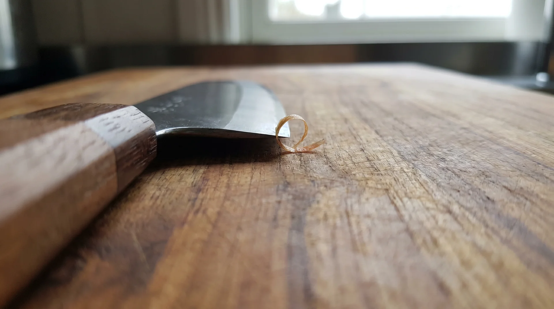 Japanese chef's knife blade in contact with teak cutting board surface, low-angle close-up