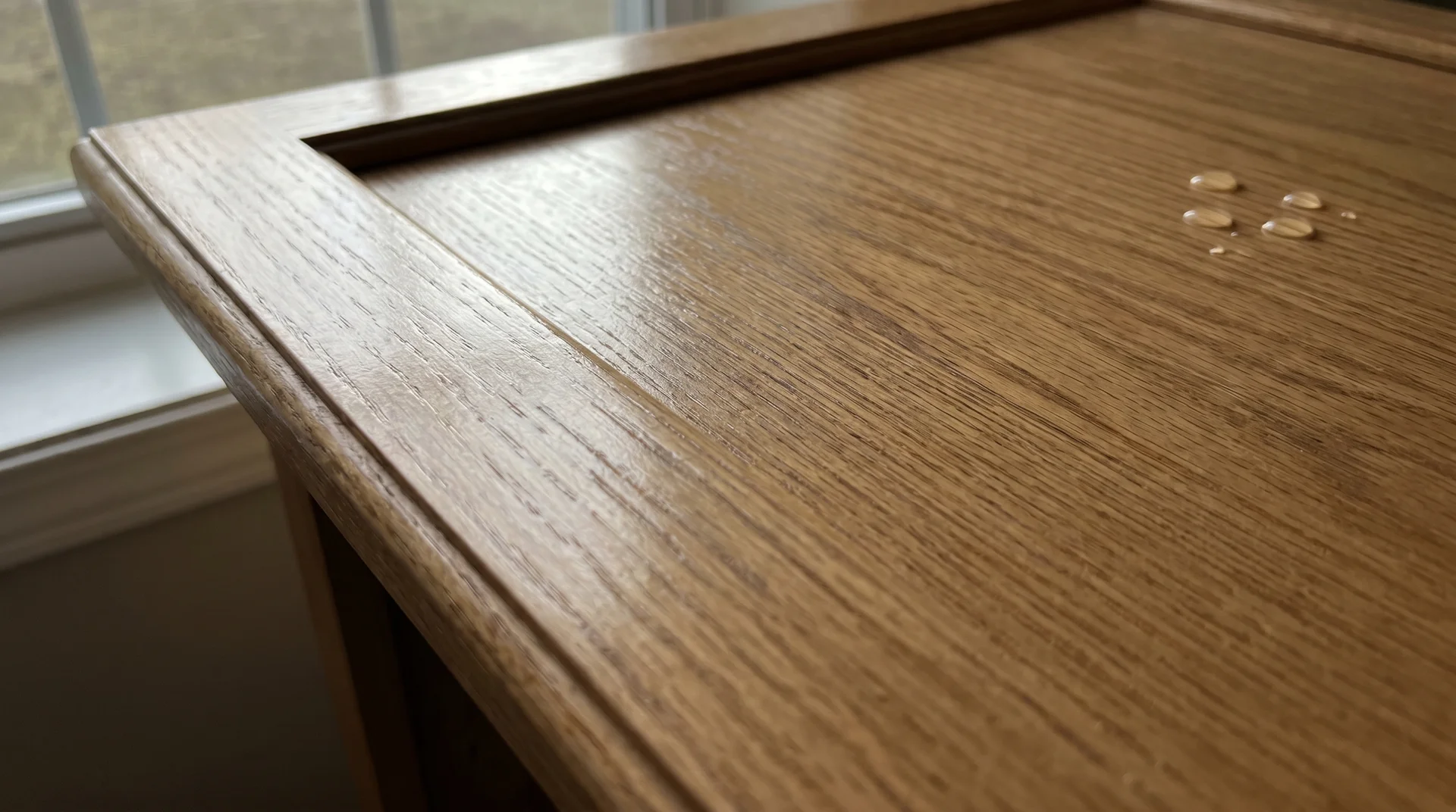 Raking light on a polyurethane-finished oak cabinet door showing surface sheen and water beading on an intact topcoat