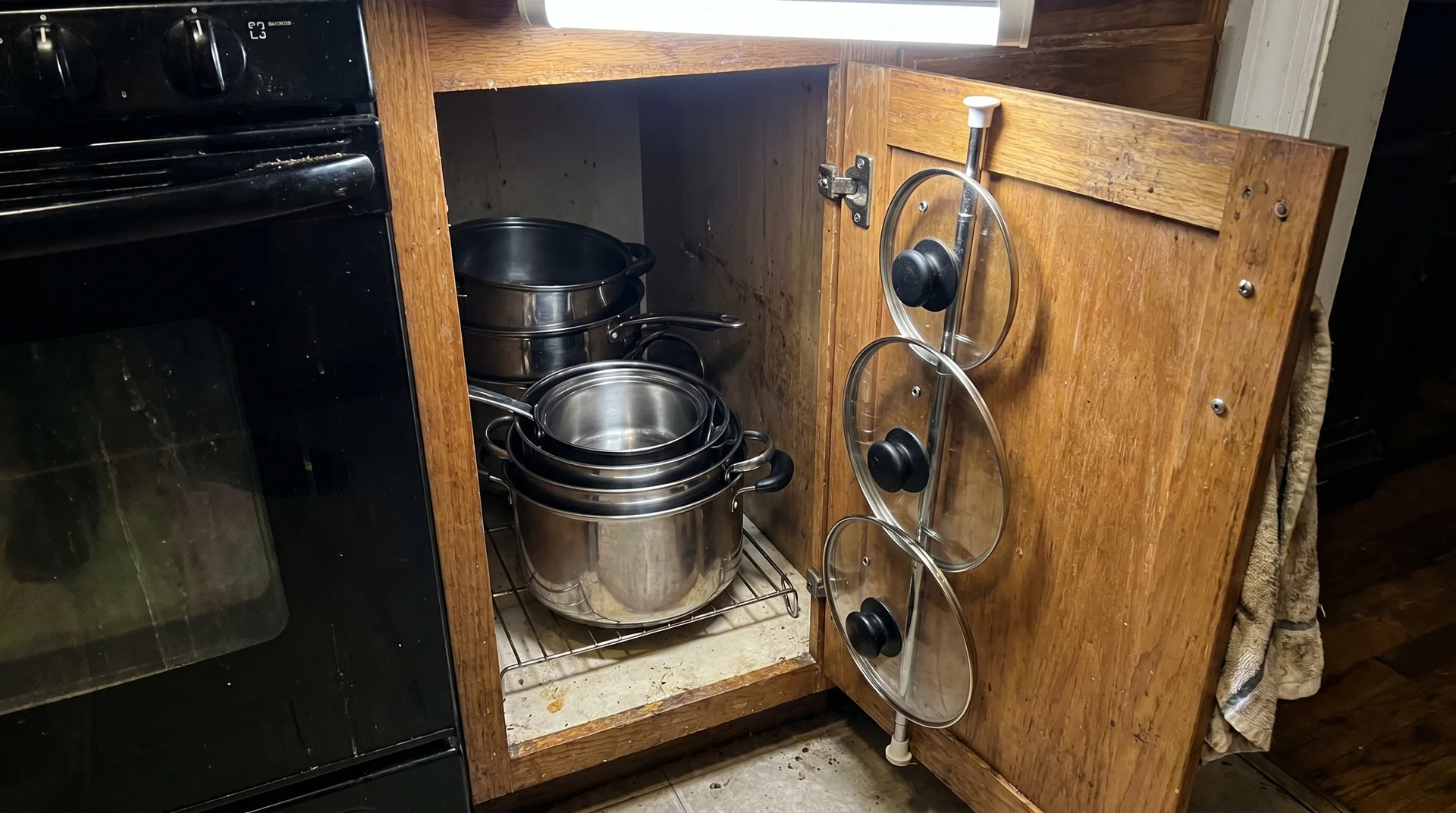 Tension rod mounted inside cabinet door holding pot lids vertically, pots nested by size below
