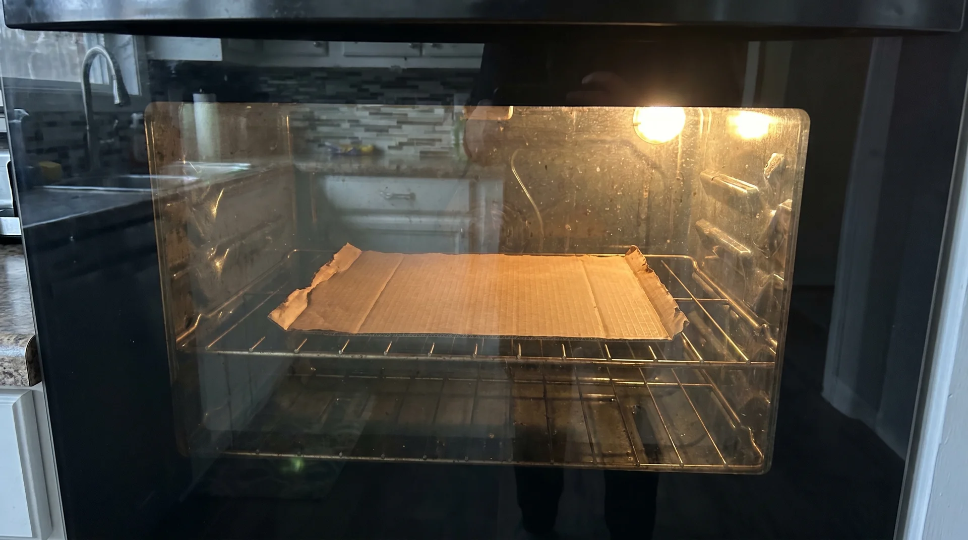 View through closed oven window showing scorched cardboard inside with oven light on