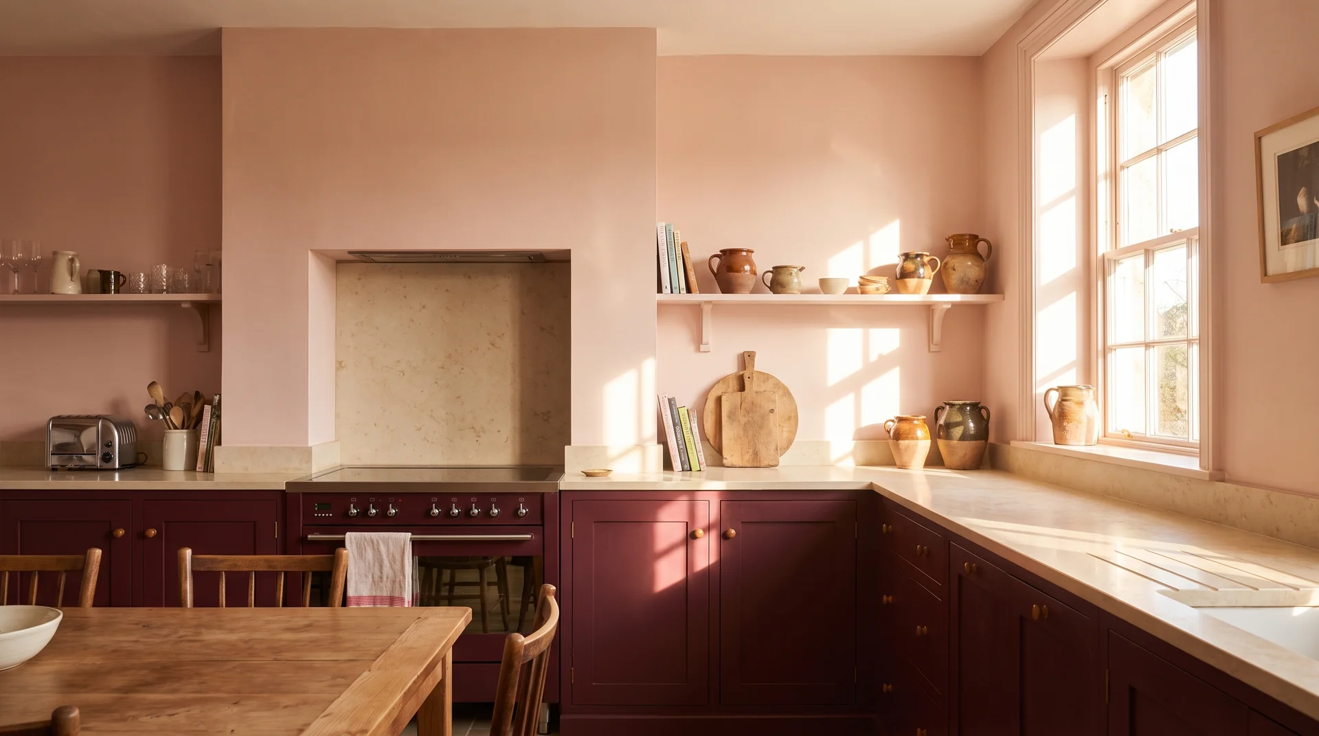 Blush pink kitchen walls beside burgundy lower cabinets in strong natural south-facing light