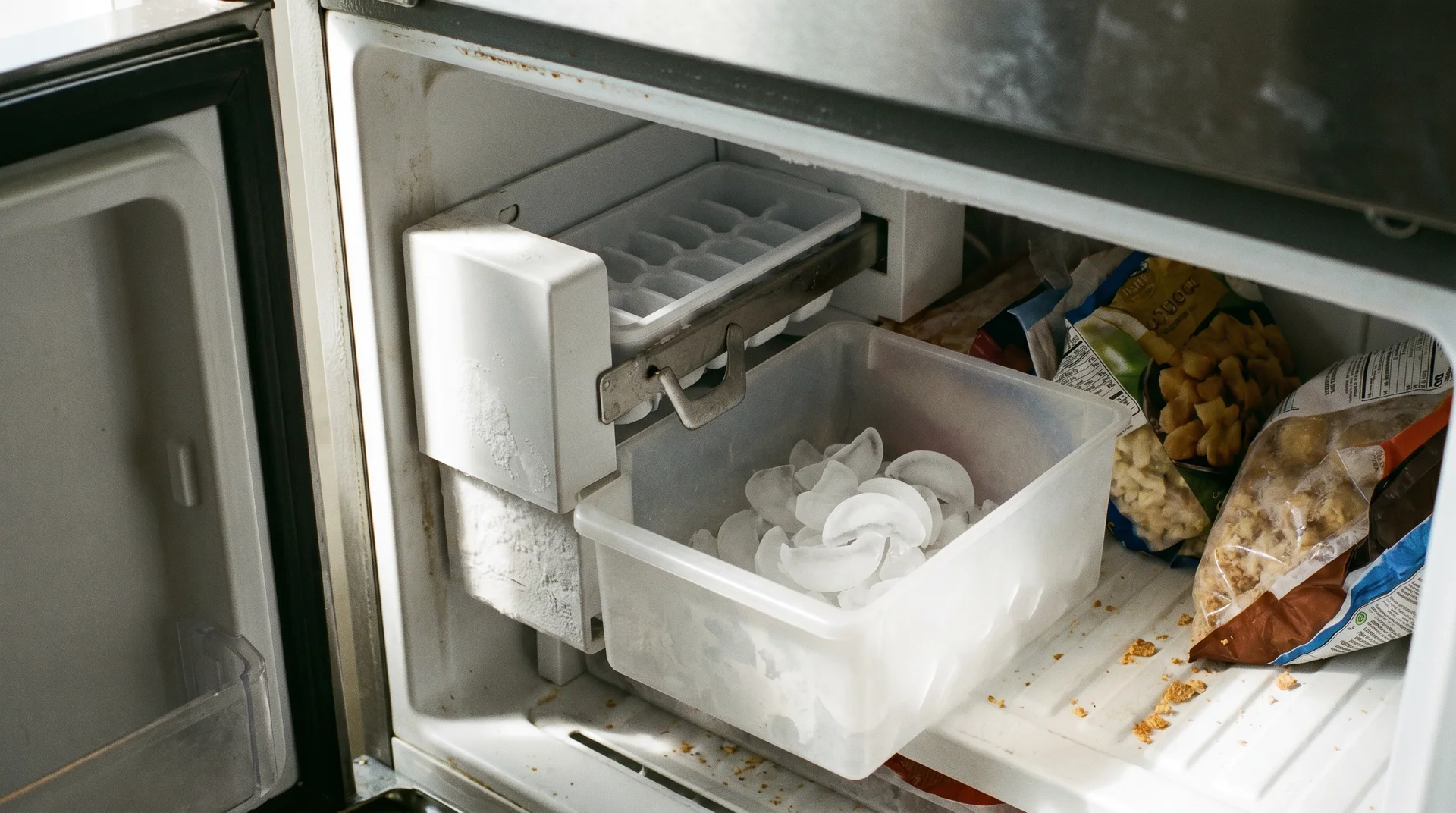 Built-in refrigerator ice maker showing tray, ejector arm, and ice bin in a residential freezer
