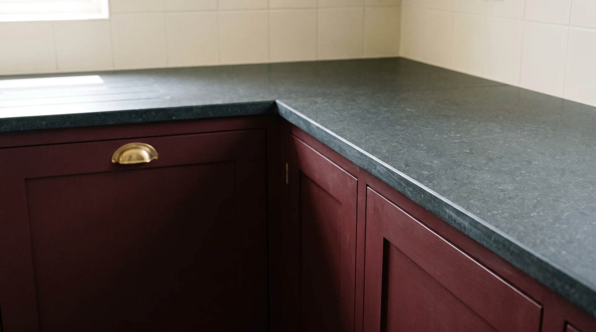 Burgundy lower cabinet below a honed charcoal quartz countertop with a brass pull and plain white backsplash