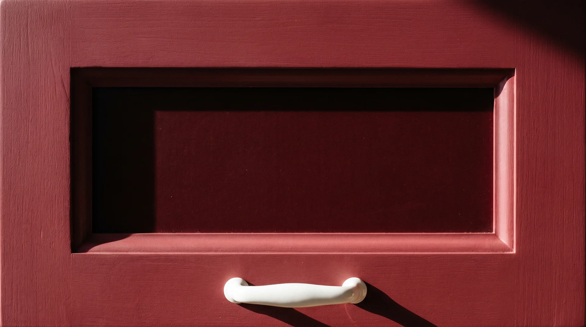 Close-up of a Shaker-profile burgundy cabinet door with ceramic pull and raking light showing panel shadow