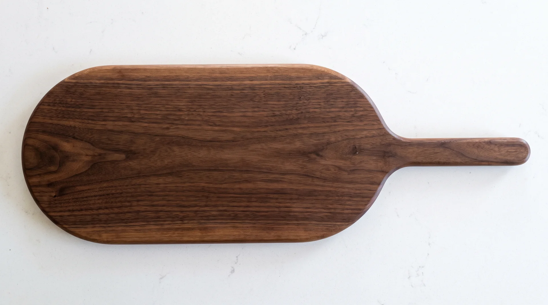 Dark walnut long paddle serving board with integral handle showing smooth roundover edges on white counter