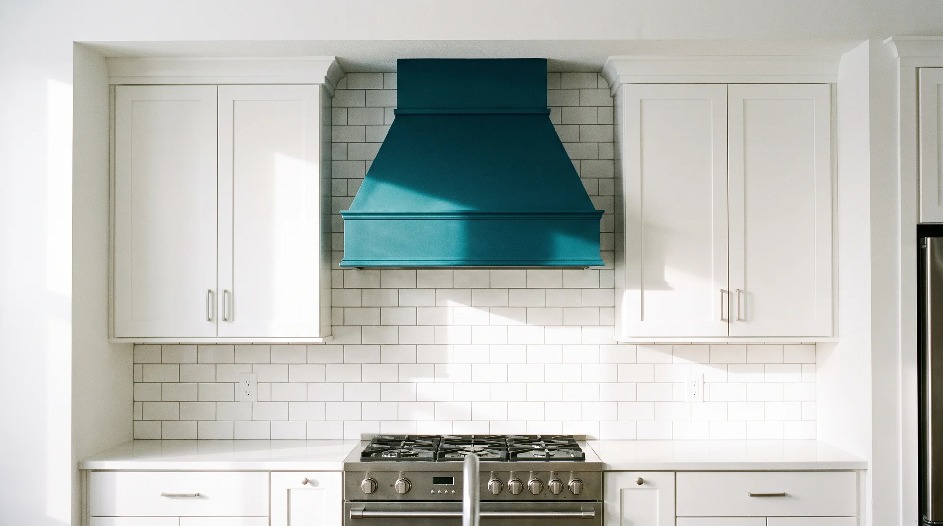 Deep teal painted range hood above a gas range with white Shaker cabinets and white tile on both sides