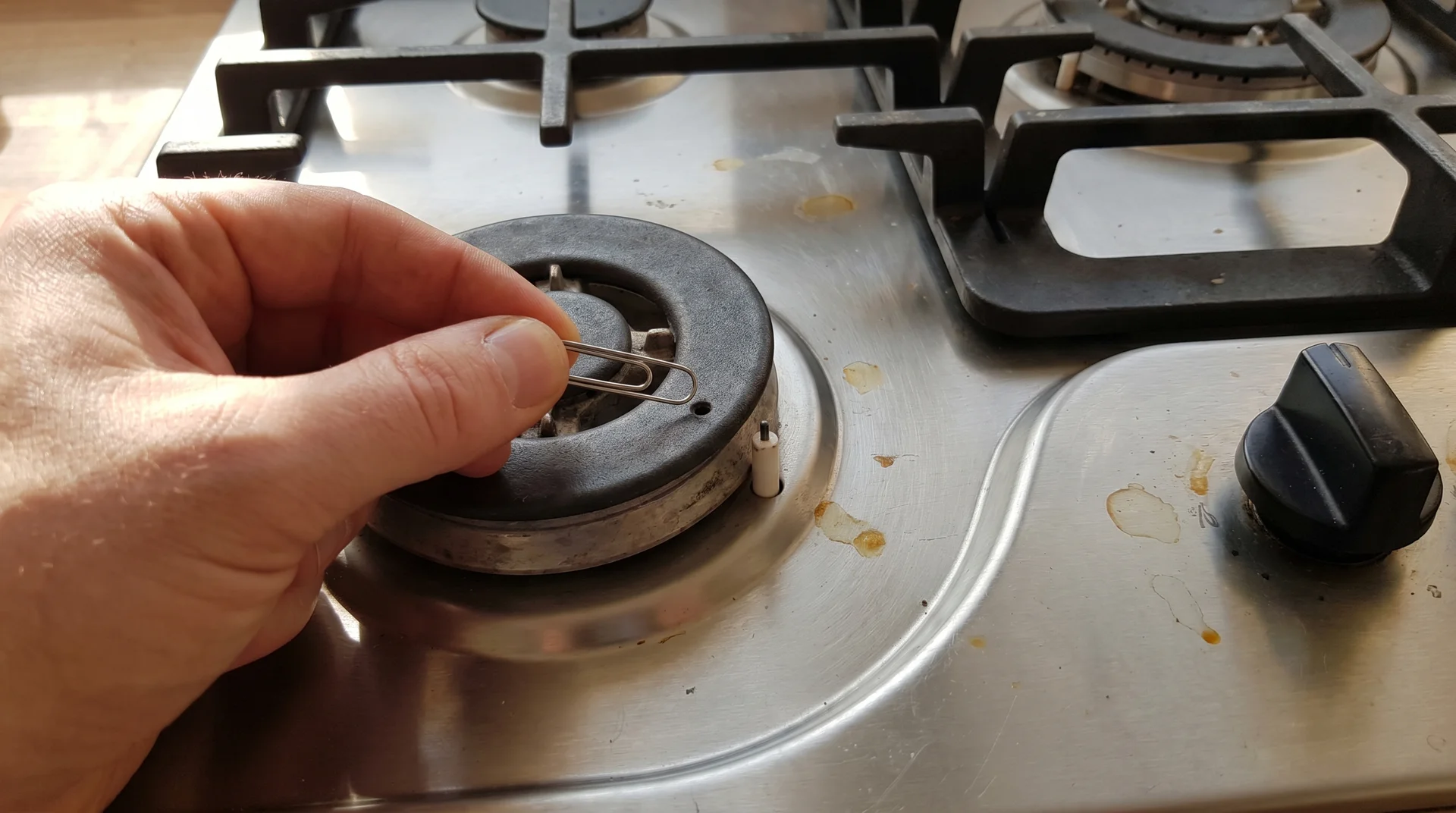 Hand clearing a gas stove burner port with a straightened paperclip, ceramic igniter tip visible beside empty burner base
