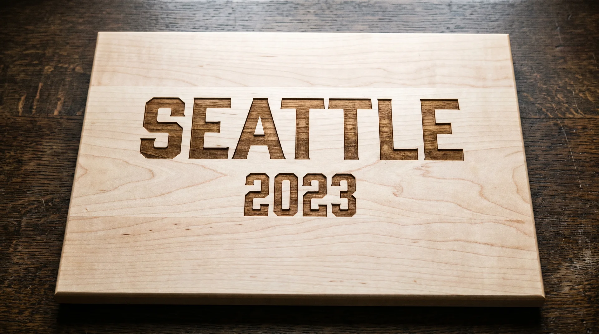 Maple board with a city name bold above and a year smaller below, viewed from above on dark oak