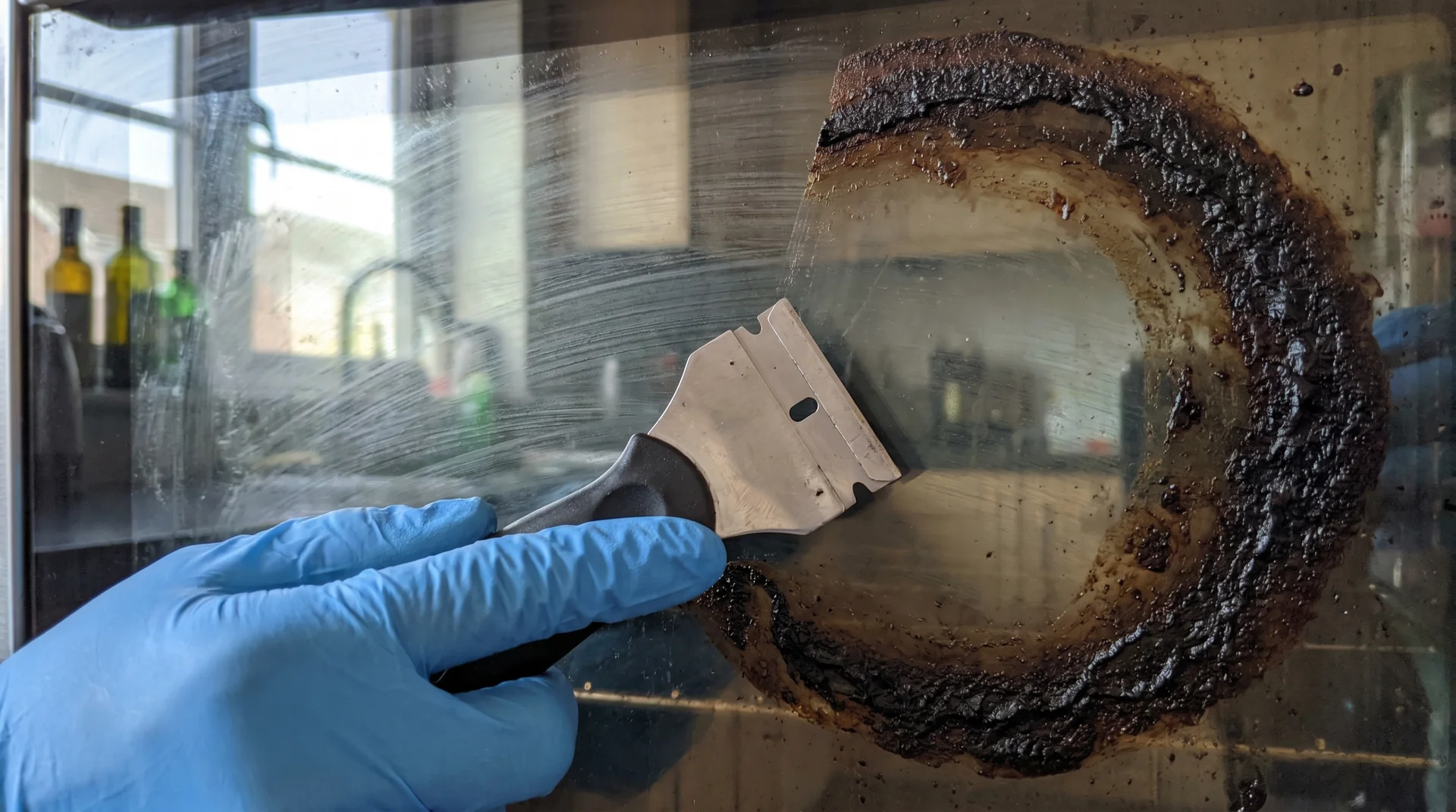 Razor blade scraper held at low 45-degree angle on oven door glass, removing carbonized residue without scratching