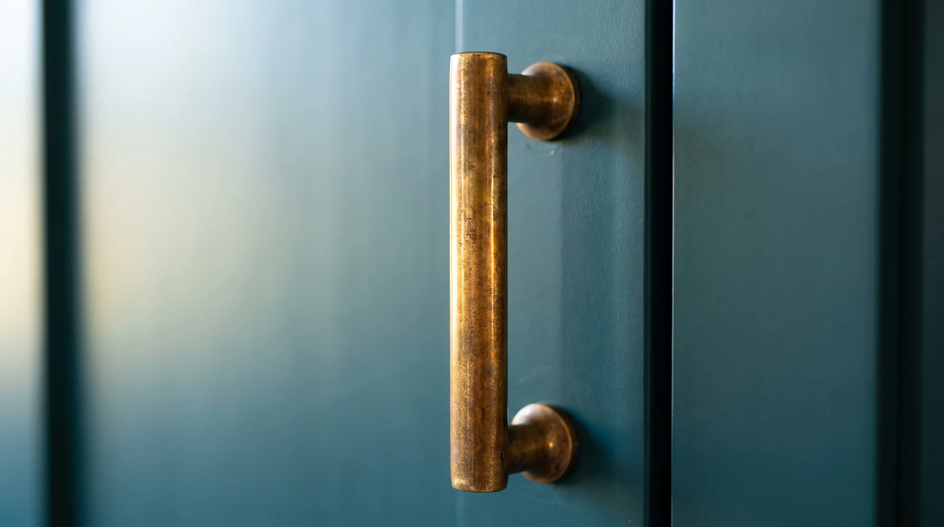 Unlacquered brass cabinet pull on a deep teal door showing the warm-to-cool contrast in natural sidelight