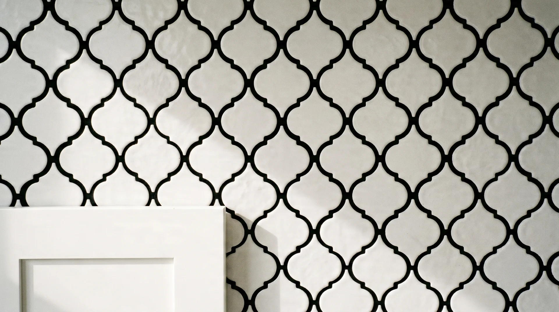 White arabesque tile with black grout close-up, tile shape outlined clearly against simple white shaker cabinet 